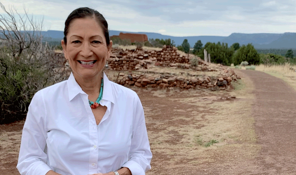 Deb Haaland smiles for the picture while standing outside in a desert terrain with short brick walls and desert brush visible behind her