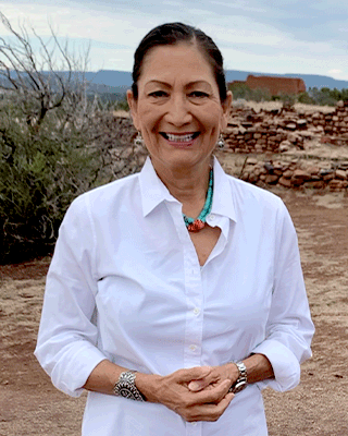 Deb Haaland smiles for the picture while standing outside in a desert terrain with short brick walls and desert brush visible behind her