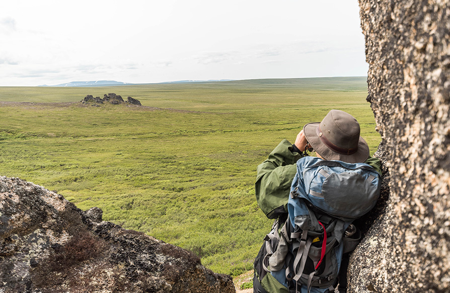 A hiker leans against a boulder while looking at a large open field through binoculars