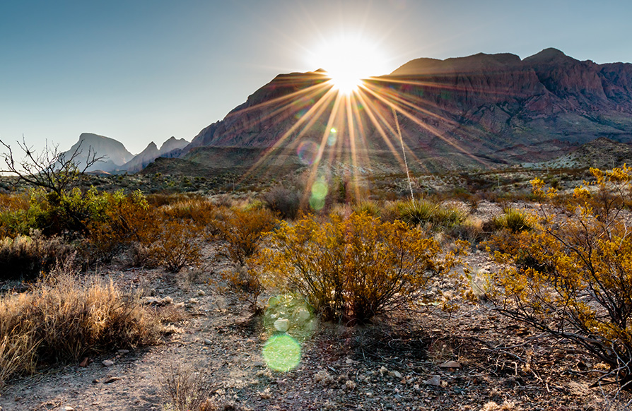The sun flares from just above a mountain peak that leads down to open land dotted with desert bushes in Big Bend National Park