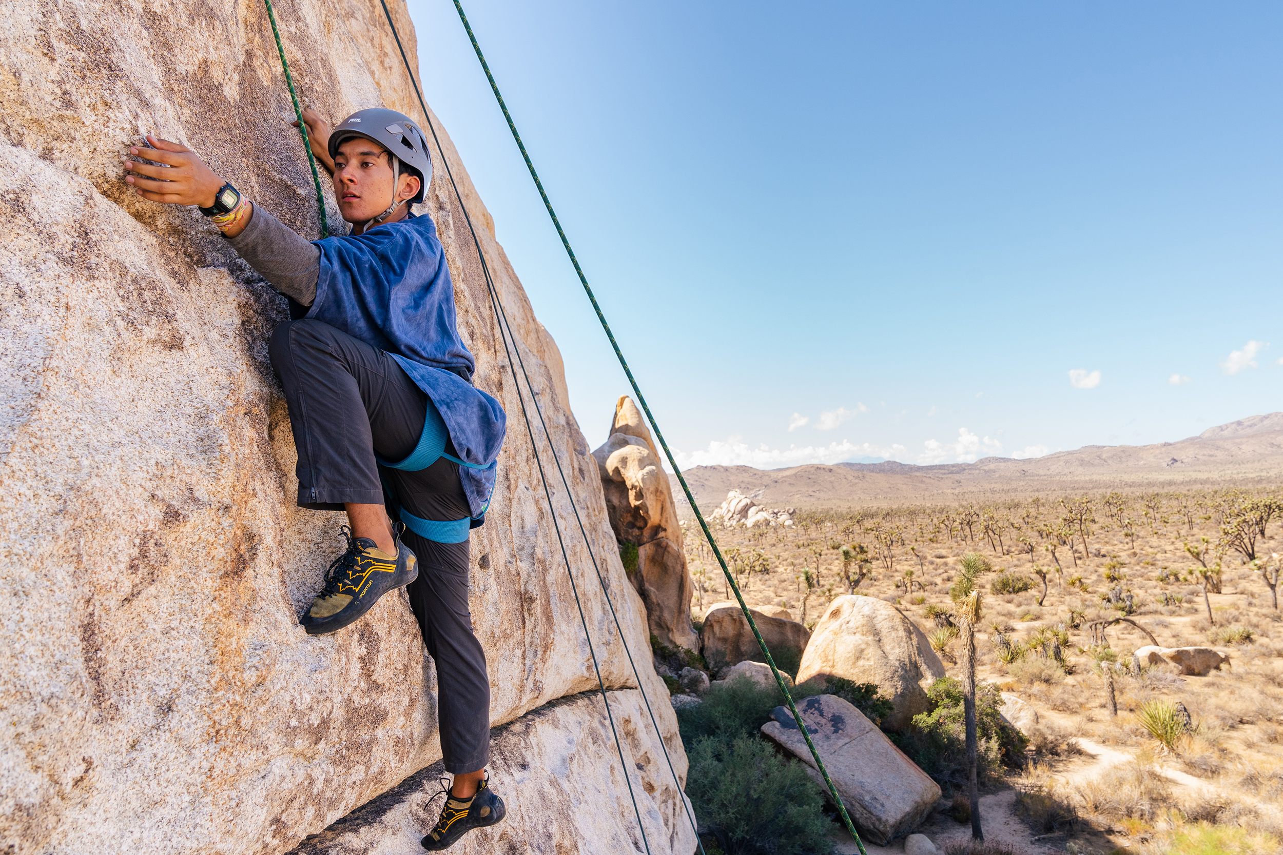 A rock climber wears a helmet and is hooked onto ropes while he scales a boulder in Joshua Tree National Park